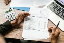Mãos seguram documentos financeiros ao lado de uma calculadora e um laptop sobre uma mesa de escritório.