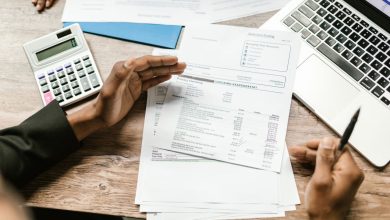 Mãos seguram documentos financeiros ao lado de uma calculadora e um laptop sobre uma mesa de escritório.