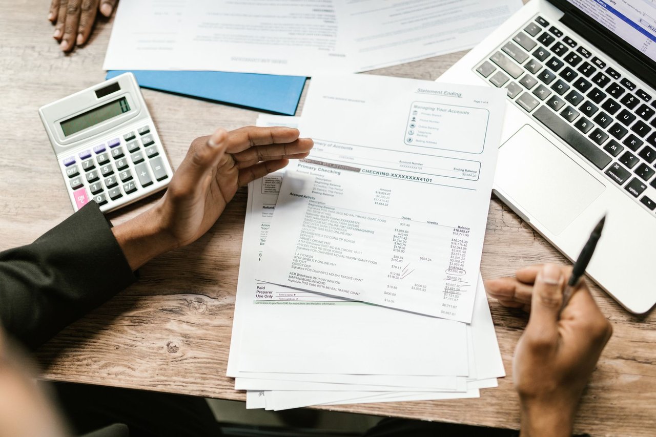Mãos seguram documentos financeiros ao lado de uma calculadora e um laptop sobre uma mesa de escritório.
